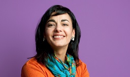 Dieses Bild zeigt Marinela Vecerik vor violettem Hintergrund. Sie hat schwarze lange Haare und trägt eine orange Weste und ein buntes Halstuch.
